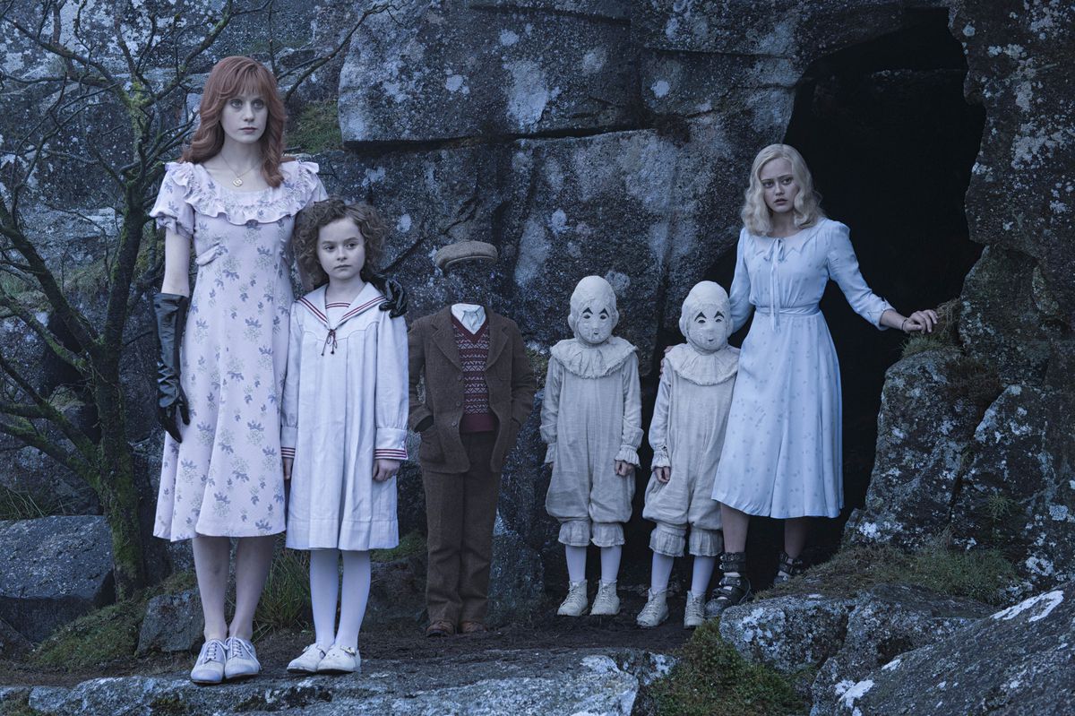 A teen with red hair and black gloves, a little curly haired brunette in a sailor’s outfit, an invisible boy dressed in a newsie outfit, two terrifying masked kids dressed in all white, and a blonde twentysomething wearing a blue dress stand in front of a cave opening in Miss Peregrine’s Home for Peculiar Children 