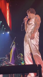 A male pop singer stands onstage beside a non-binary pride flag.