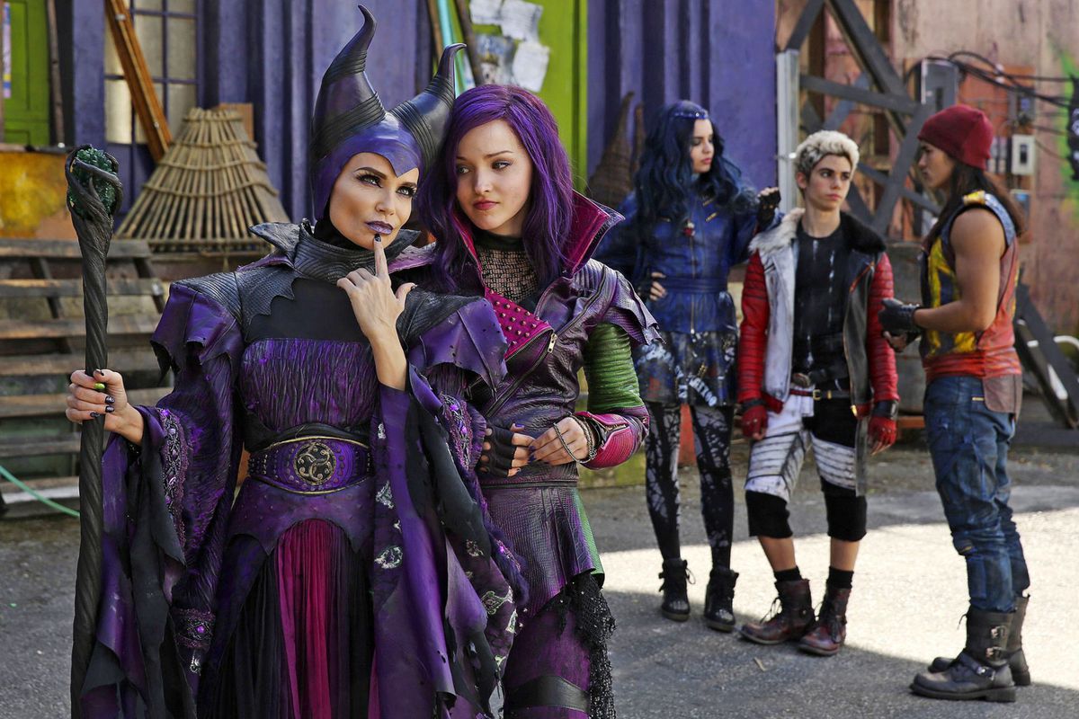 A woman in a purple witch costume with horns stands next to a girl with purple hair in a leather biker jacket with three other kids in the background wearing costumes.