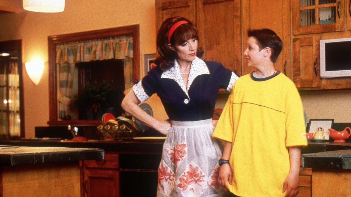 A brown haired woman in a navy blue maid outfit with a flower apron and red headband stands next to a teenage boy in an oversized yellow shirt with her hands on her waist in a kitchen.