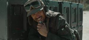 Ruescott Melshi holds a transmitter close to his mouth while wearing his signature floppy hat and glasses in Rogue One.
