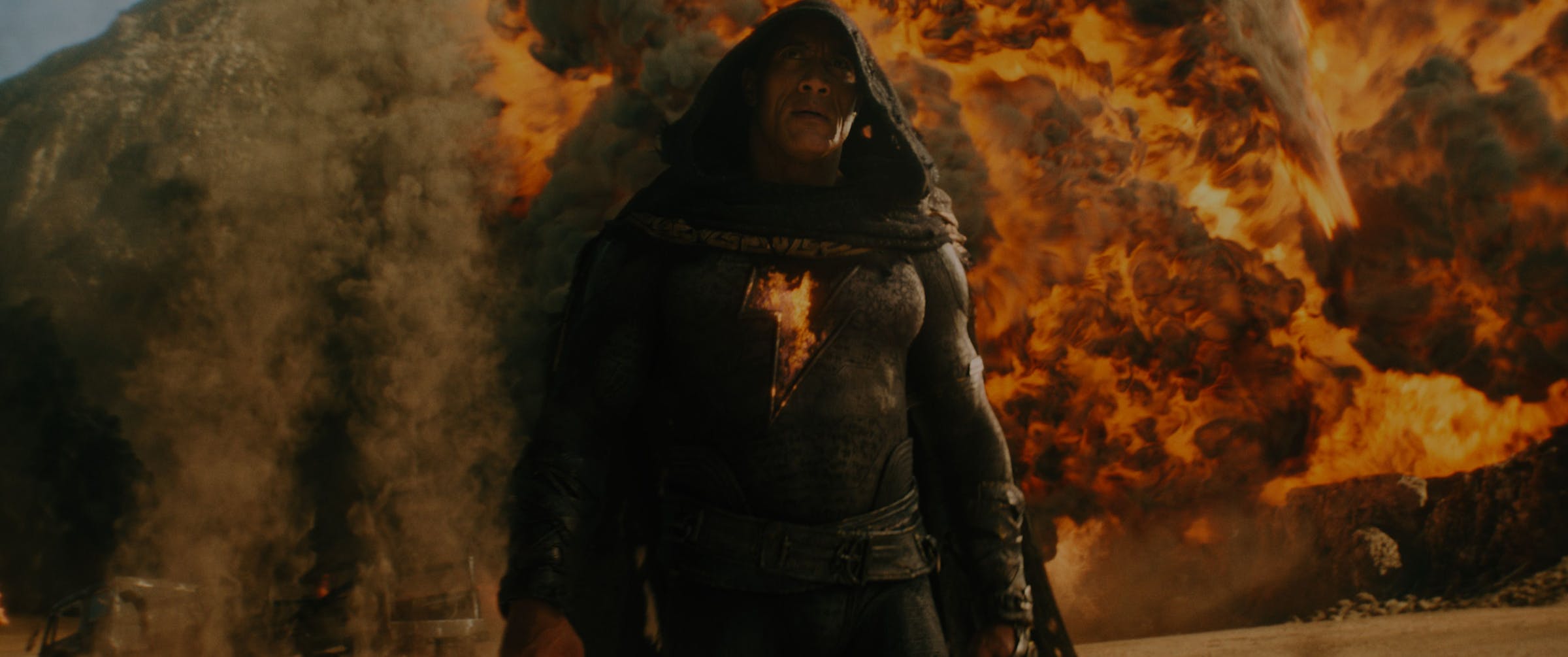 A man in a black body suit emblazoned with a glowing lightning bolt and accented by a black hooded cape. Behind the man is a wall of flames from an explosion he’s caused.