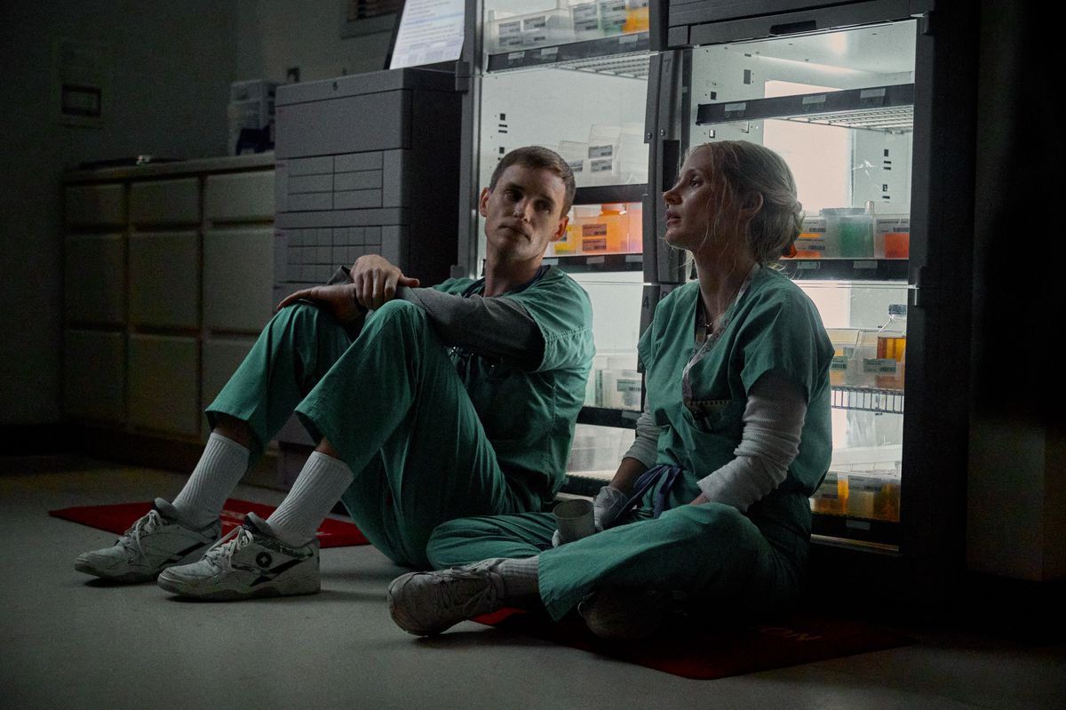 Two nurses, male and female, in green scrub outfits sitting on the floor next to a glass freezer full of vials, looking exhausted.