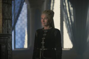 Eve Best, wearing formal clothes with her hair up, stands in front of open curtains in House of the Dragon.