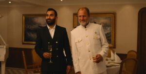 The unnamed yacht captain (Woody Harrelson) in Triangle of Sadness, in full white dress uniform, stands next to a black-clad, smiling guest on the yacht at the Captain’s Dinner