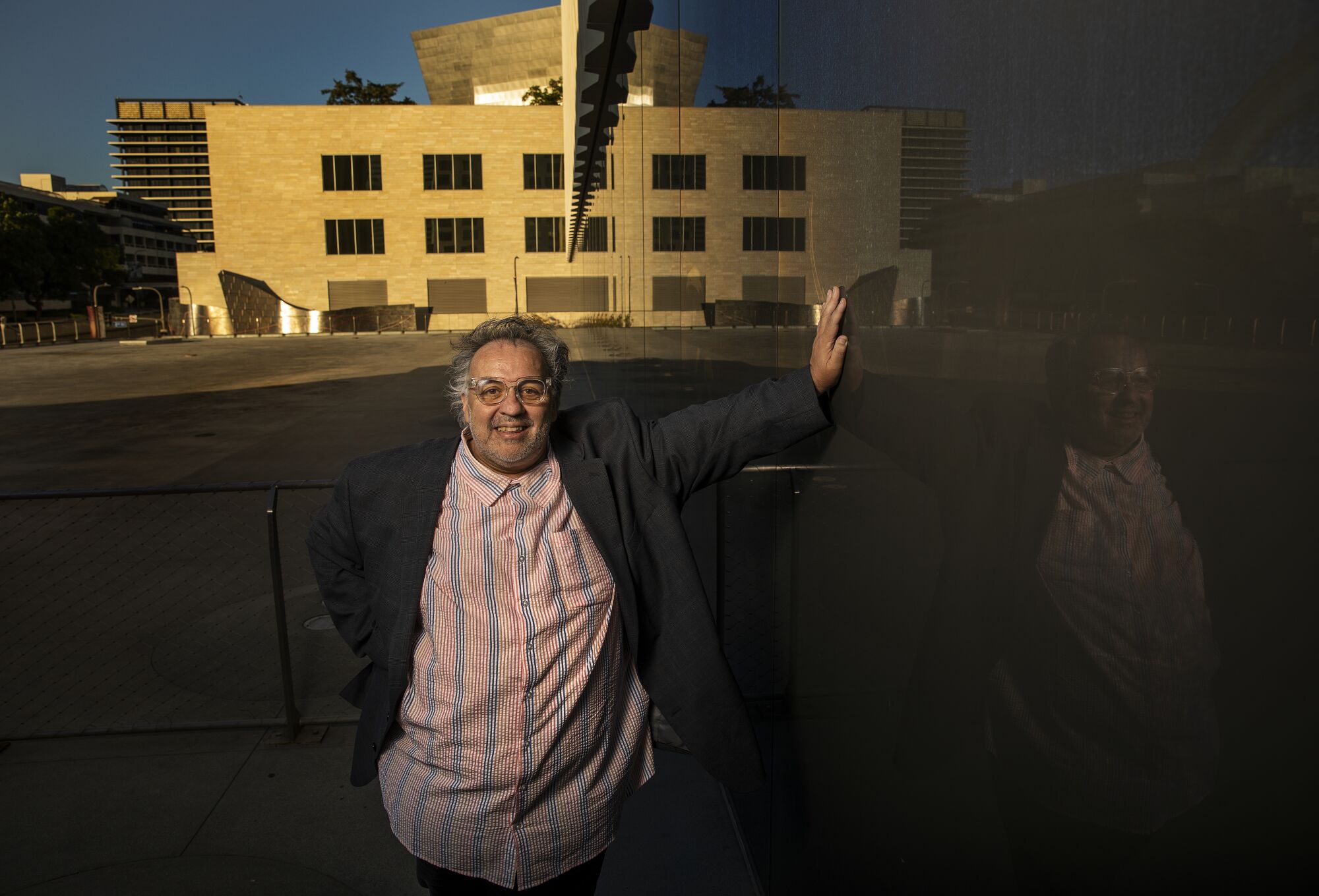 Luis Alfaro, in a dark jacket and a striped shirt, leans against a glassy wall in afternoon light