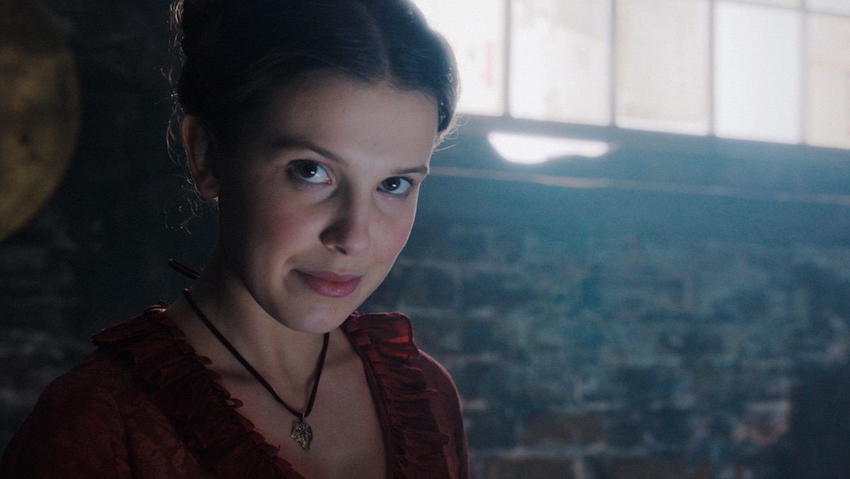 Millie Bobby Brown in a red dress as Enola Holmes smirks at the camera in front of a window in a stone wall