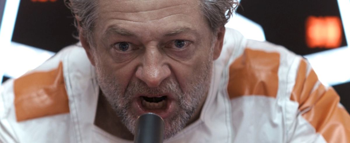 Kino Loy (Andy Serkis), still in his white and orange trimmed prison jumpsuit, yells into a microphone commanding his fellow prisoners to leave