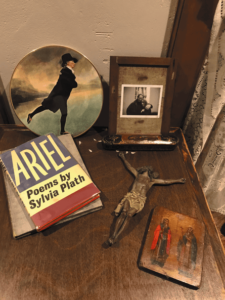 Photograph of a wooden table on which there are various objects: a display plate of the painting he Skating Minister by Henry Raeburn’ a framed photograph, a crucifix, a small painted icon and a hardback copy of Ariel by Sylvia Plath