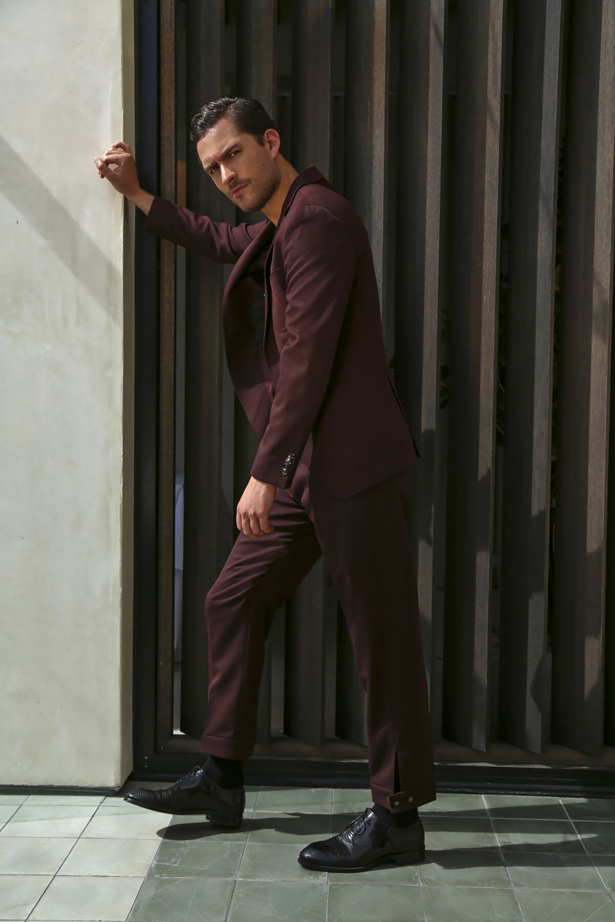 A man in a plum-colored suit stands leaning with one hand against a wall.