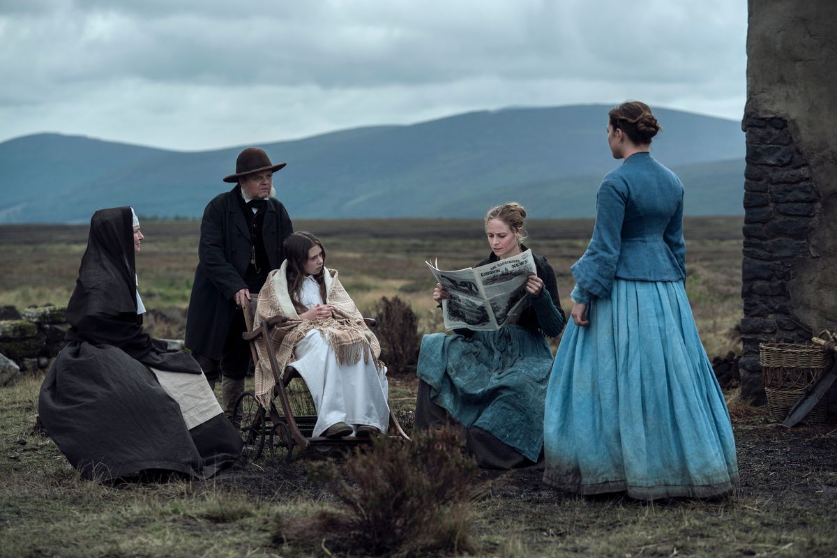 A nun (Josie Walker), a bowler-hatted doctor in all black (Toby Jones), a girl wearing white and sitting in a wheelchair (Kíla Lord Cassidy) and two women in long blue skirts (Niamh Algar and Florence Pugh) sit outside on a featureless plain near a crumbling building in The Wonder