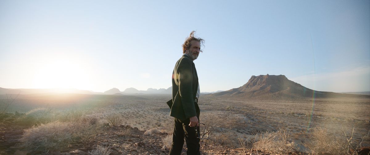 Silverio (Daniel Gimènez Cacho), a shaggy-haired man in a plain black suit, stands in a sun-blasted desert with a mesa in the far distance, looking back over his shoulder at the camera, in Bardo: False Chronicle of a Handful of Truths