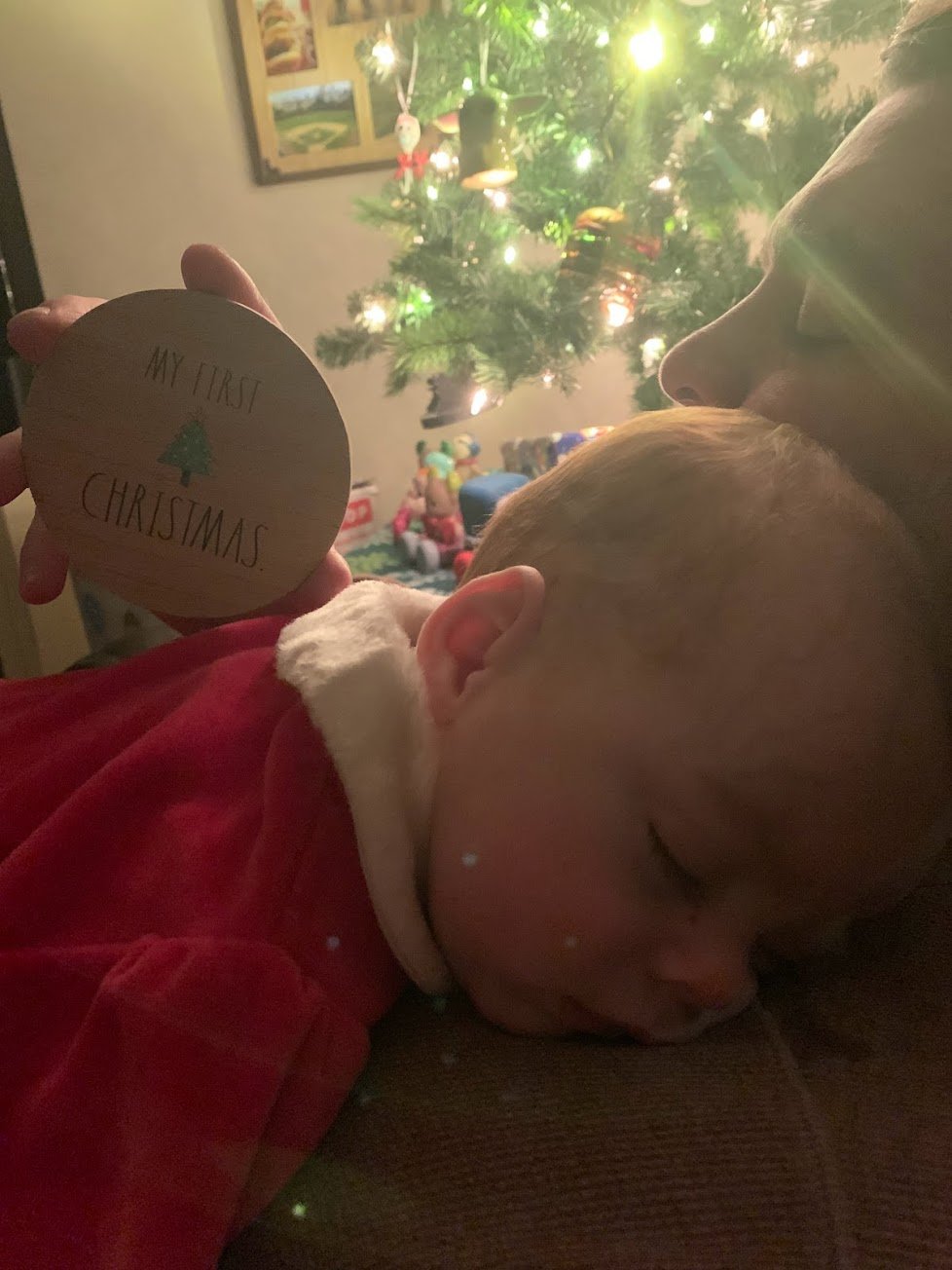 A man holds his sleeping son in front of a Christmas tree while holding a sign that says My first Christmas