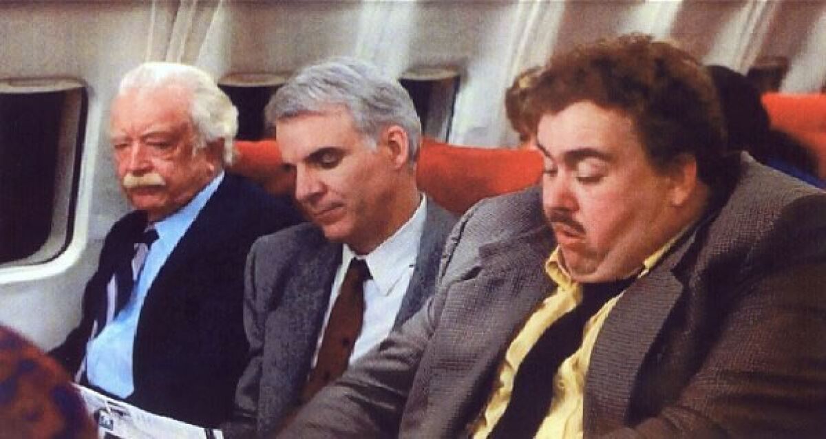 Three men seated in a commercial airplane in the movie "Planes, Trains, and Automobiles."