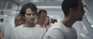A medium shot of a white hallway filled with men in white jumpsuits accented with orange pads on their left shoulders.