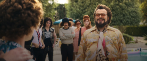 Musician Wolfman Jack (played by Jack Black) smirks at “Weird Al” Yankovic (Daniel Radcliffe) by a backyard pool at the home of Dr. Demento (Rainn Wilson), as ’80s celebrities including Gallagher (Paul F. Tompkins) and Alice Cooper (Akiva Schaffer) look on in Weird: The Al Yankovic Story