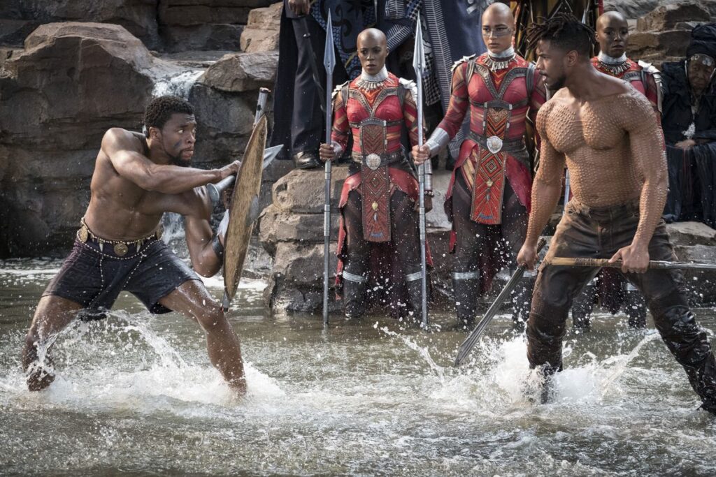 T’Challa/Black Panther (Chadwick Boseman) and Erik Killmonger (Michael B. Jordan) fight shirtless in ankle-deep water against a background of Dora Milaje observers in 2018’s Black Panther