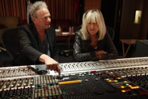 Man in black blazer and woman in black leather jacket sit in front of music studio sound mixing board.