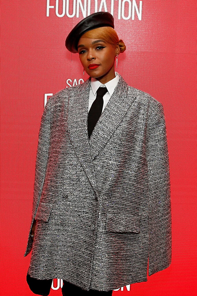Janelle Monae smiles in gray suit and black tie against red backdrop