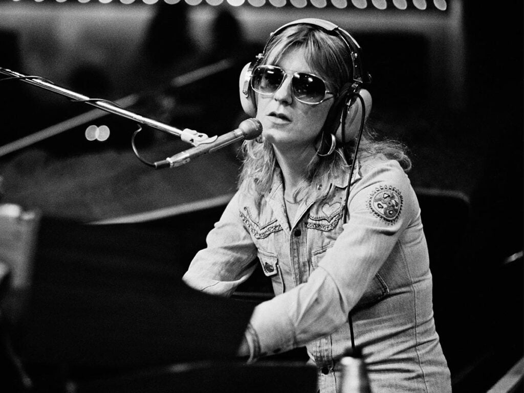 Black and white photo of Christine McVie sitting at an organ wearing sunglasses in the 70s
