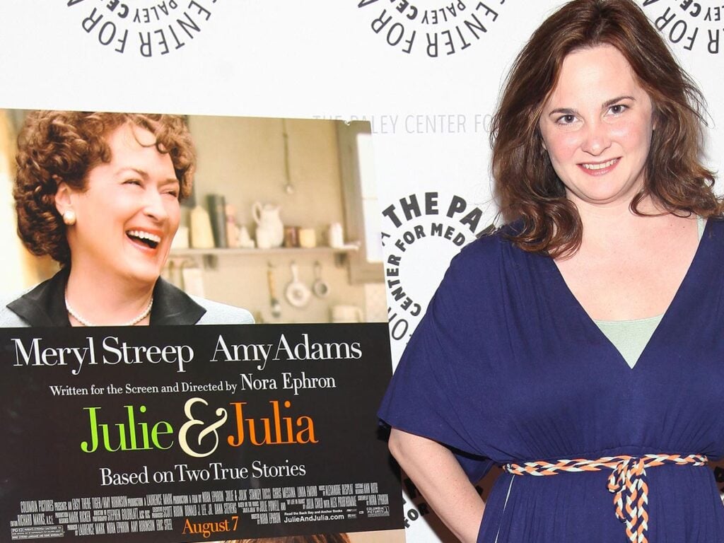 Julie Powell in blue standing next to a poster for the movie Julie and Julia