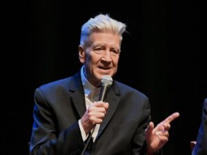 David Lynch in a black suit jacket and white shirt talking into a microphone on a darkened stage