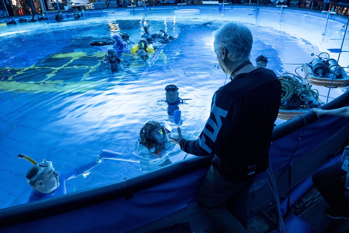 James Cameron, back turned to camera, speaks to Spider actor Jack Champion who is wearing his prop facemask and floating in the water next to two crew members with snorkel masks, and a floating set behind them. They are all in a dive tank built for Avatar: The Way of Water.