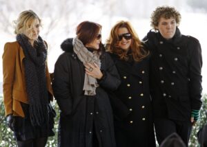 Three women bundled up in coats and a man with curly hair in a dark coat.