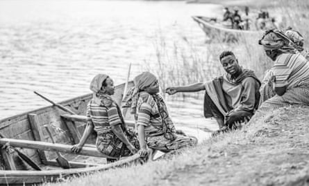 Female fishers sit in a boat and also on the bank, talking to Makembe. All wear traditional dress