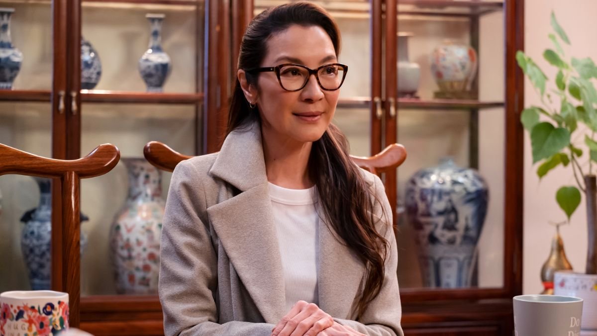 Michelle Yeoh sitting at a dining room table wearing a white shirt, tan overcoat, and black rimmed glasses