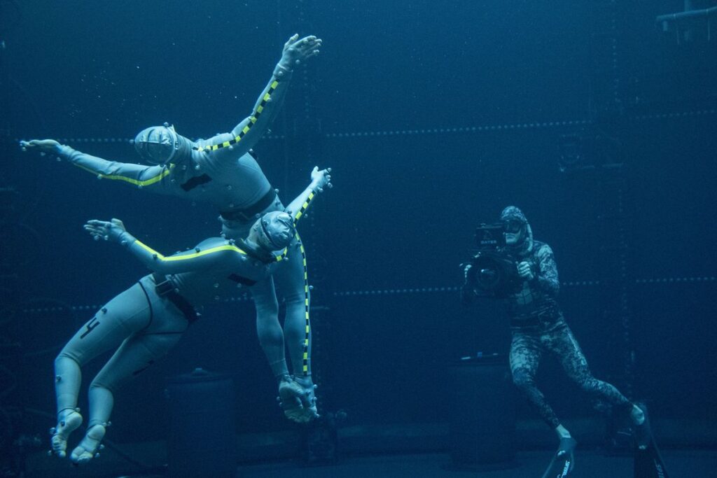 An underwater motion capture technician holds up a camera-like device pointed at two actors swimming around with their arms open in a pool for Avatar: The Way of Water