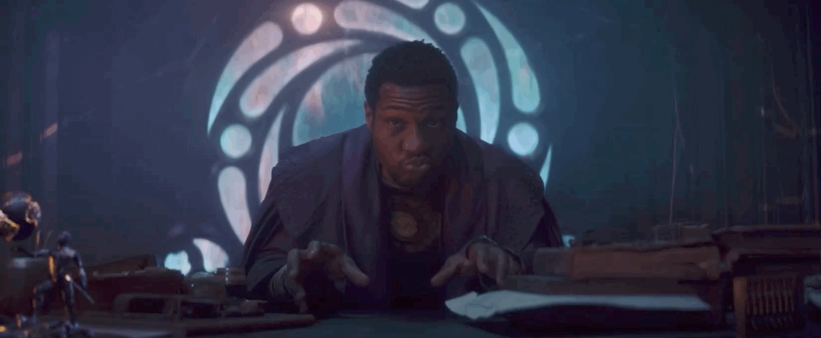 Jonathan Majors as He Who Remains sits at his desk ans mimics an explosion with his hands and mouth on Loki