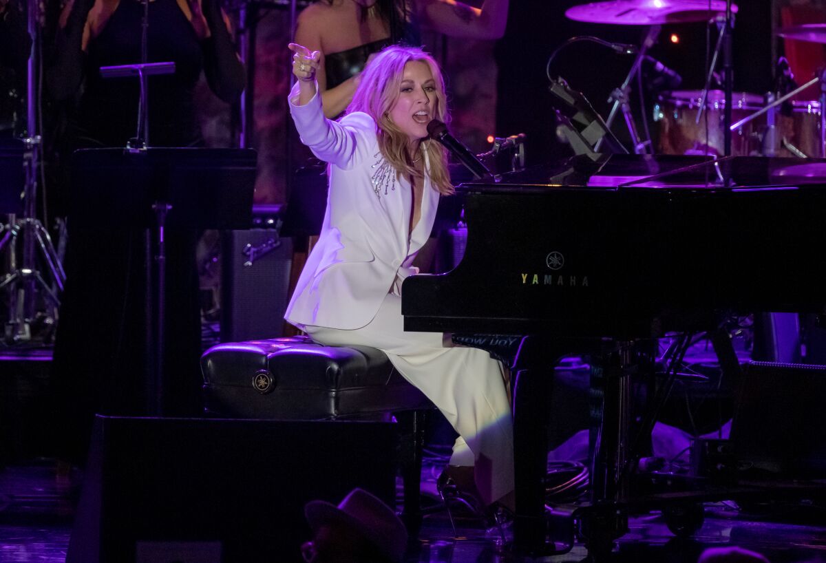 A woman in a white suit sings and plays the piano