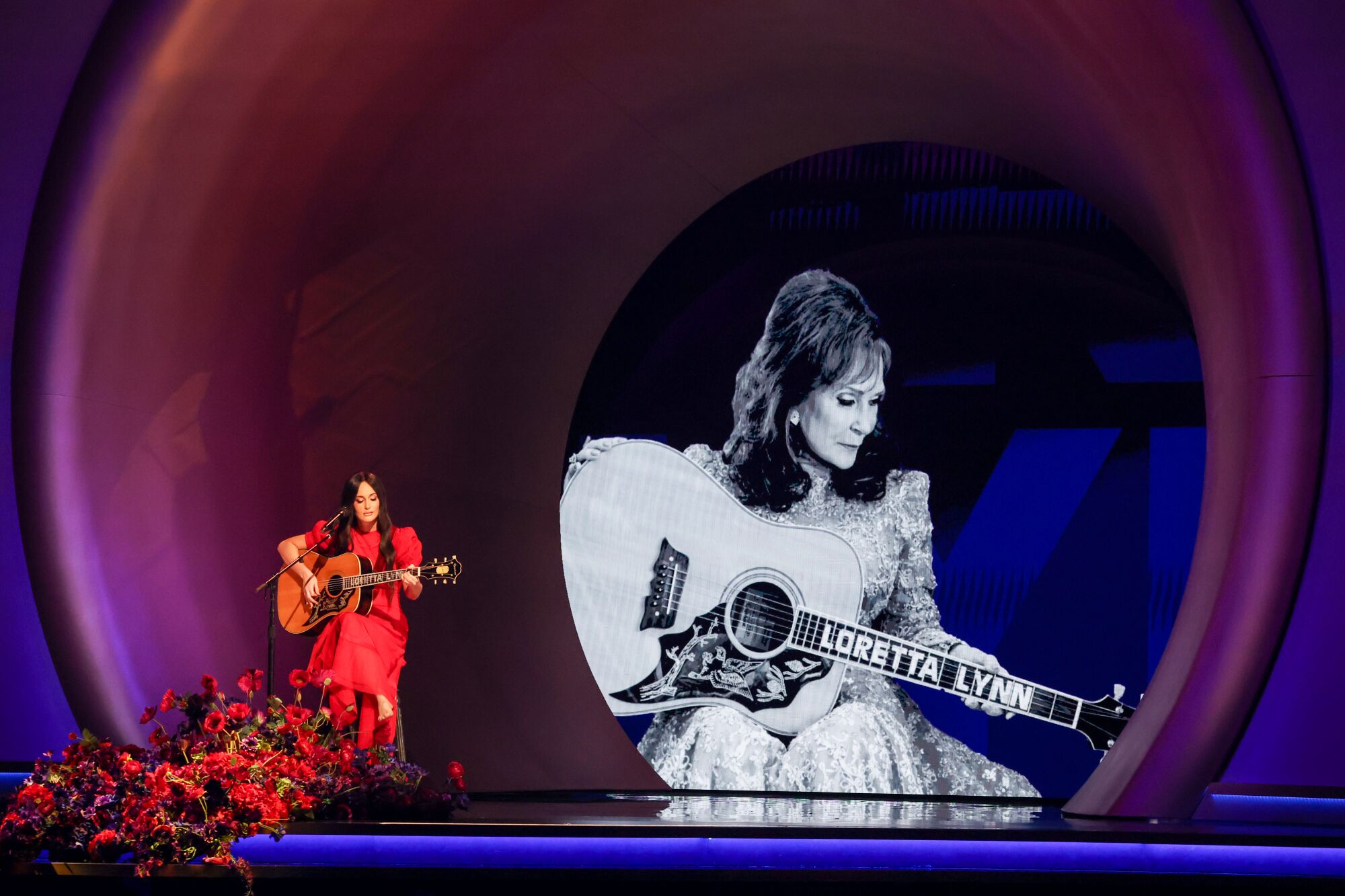 A woman sitting and playing guitar in front of a mic and giant black-and-white image of a woman playing guitar