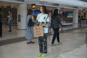Storm Reid, wearing the pajamas + crocs fit one wears to the airport, holds a cardboard sign that says “Welcome Back Mom!” in Missing.
