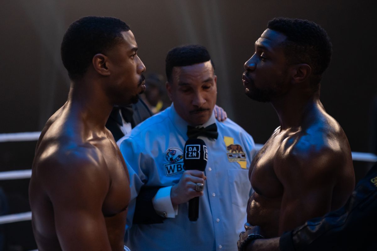 Adonis Creed (Michael B Jordan) and Damian (Jonathan Majors) stand shirtless opposite each other with a boxing ref in the middle ready to fight in Creed III