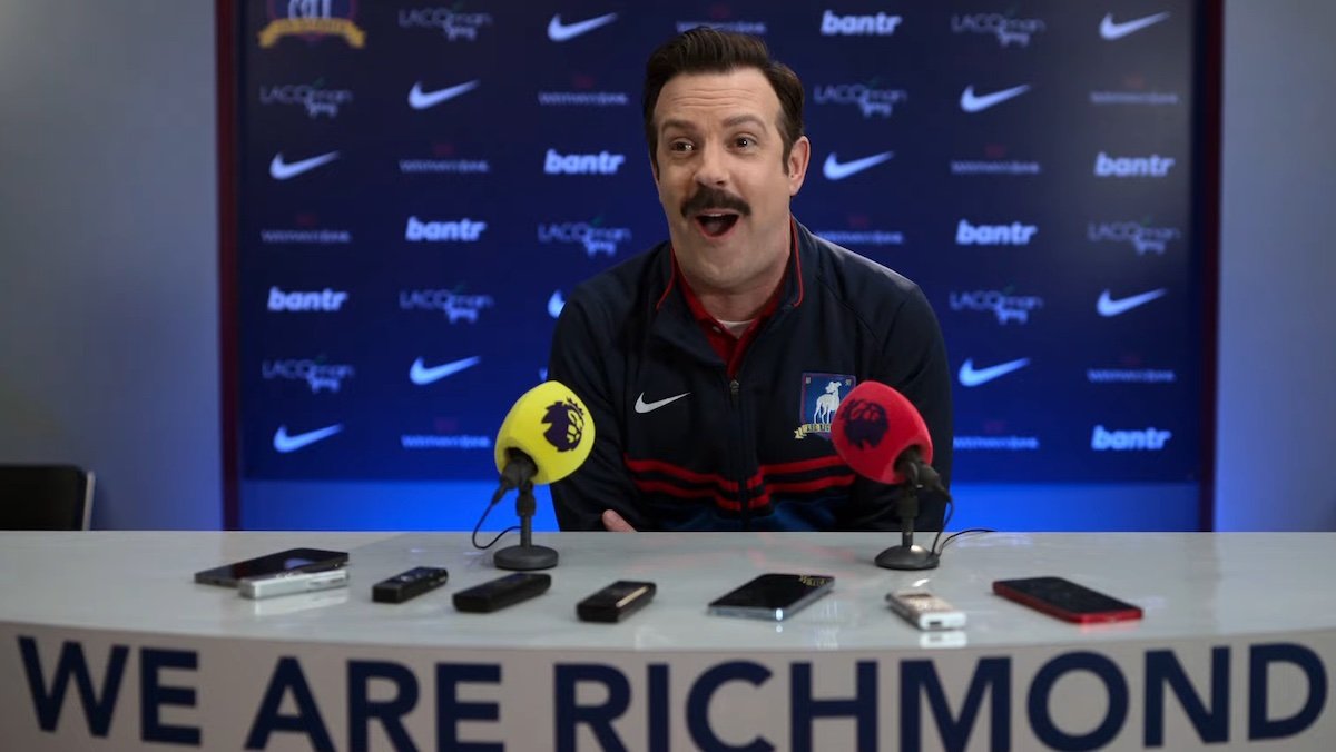 Jason Sudeikis's Ted Lasso makes a big O face behing a podium during a press conference on Ted Lasso season 3