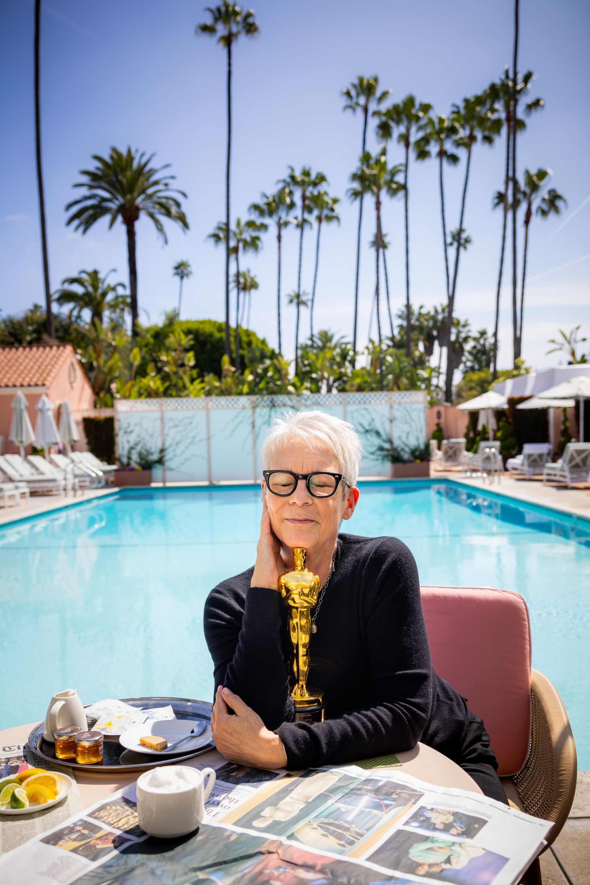 A woman in glasses and a dark sweater holds her Oscar trophy in front of a swimming pool.