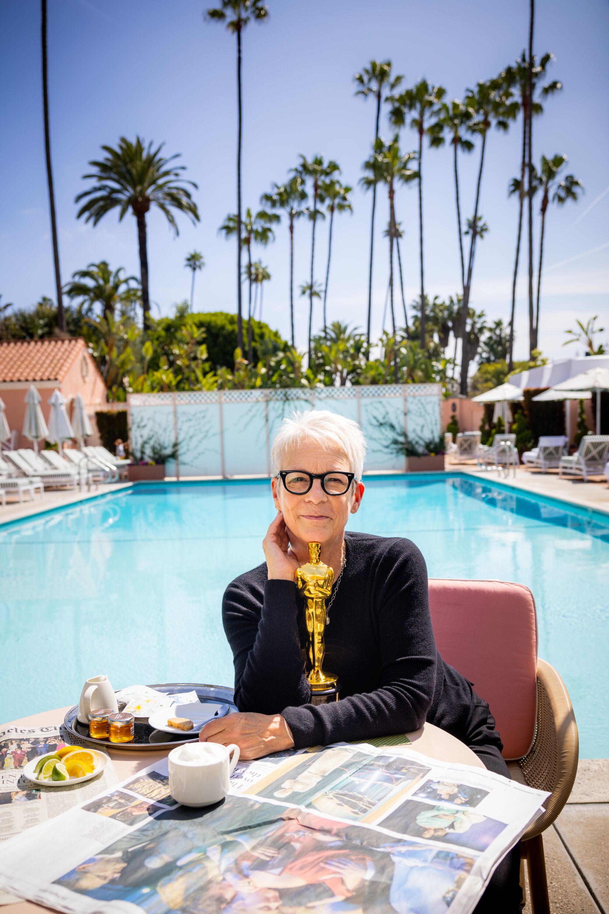 A smiling woman in glasses and a black sweater holds her Oscar trophy in front of a swimming pool.