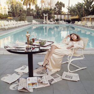 A woman in a robe lounges in front of a swimming pool, with her Oscar on a table and newspaper pages at her feet.