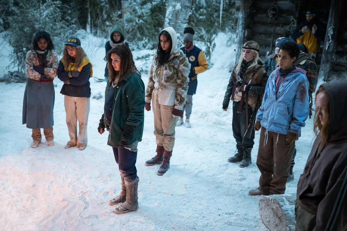 The teen Yellowjackets stand in the snow outside of their cabin, with a campfire just offscreen in the season 2 premiere of Yellowjackets.