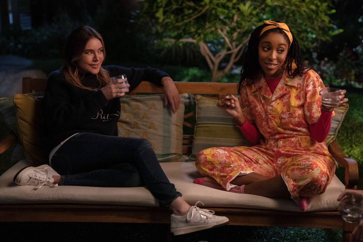 Christa Miller, in a black top and pants, and Jessica Williams, in an orange jumpsuit, sitting on a couch and holding drinks