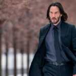 Keanu Reeves' John Wick in a black suit and long hair walks down a road lined with trees in Chapter 4