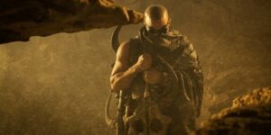 Riddick, wearing a cloak and his goggles, carrying the tail of an alien monster.