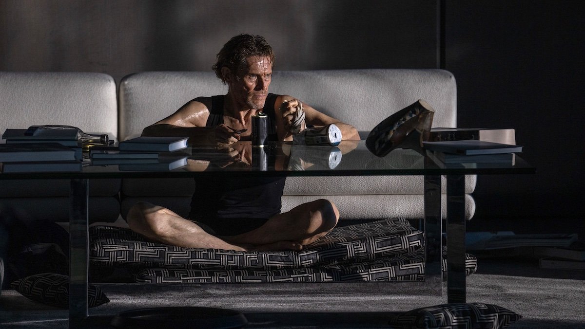 Willem Dafoe in a blank tank top sits on the floor at a coffee table in Inside