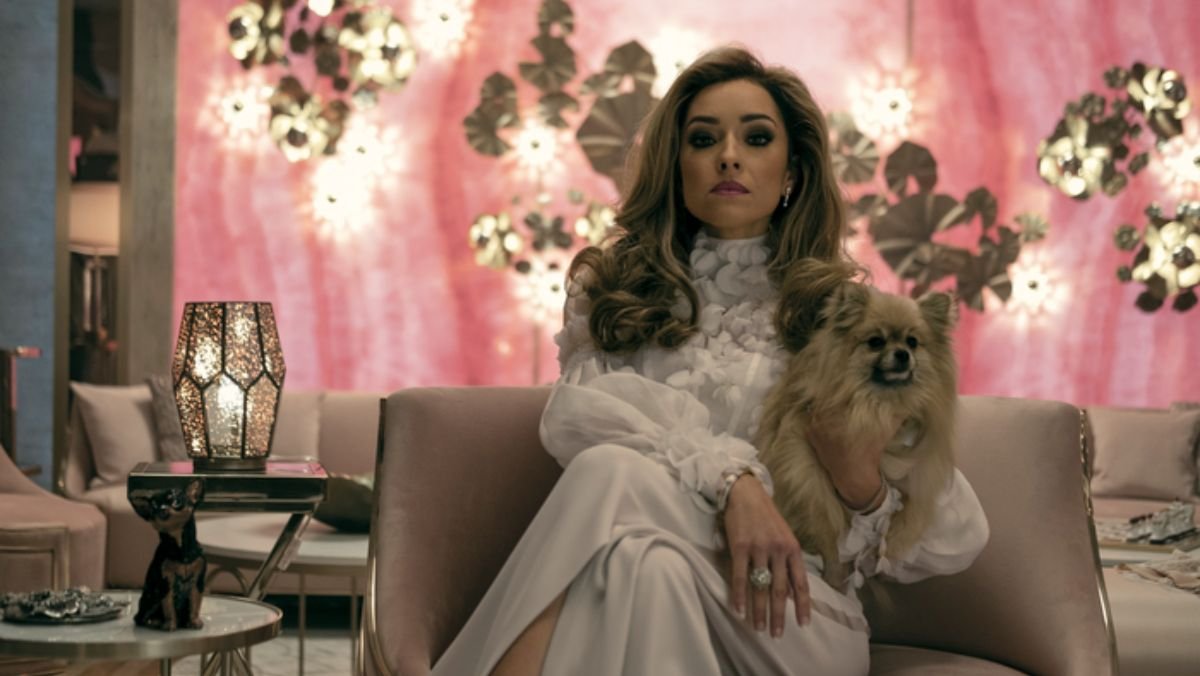 Tatiana sits in a champagne colored chair with a pink background holding a small dog in a scene from The Power featurette