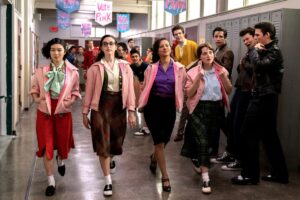 Four young women wearing pink jackets over 1950s outfits walk past leering young men in a school hallway