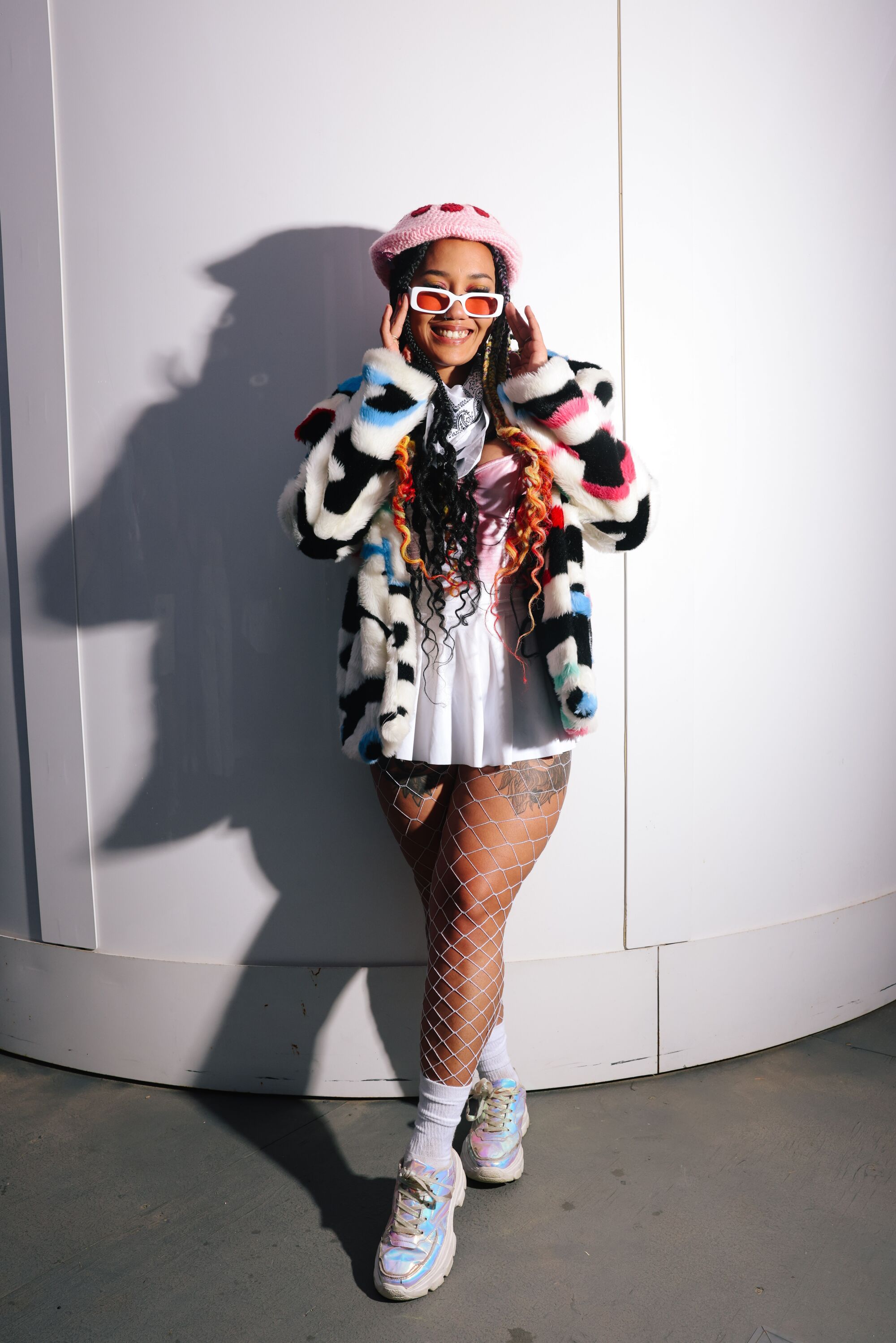 A woman wearing a hat, furry coat and sunglasses poses against a wall