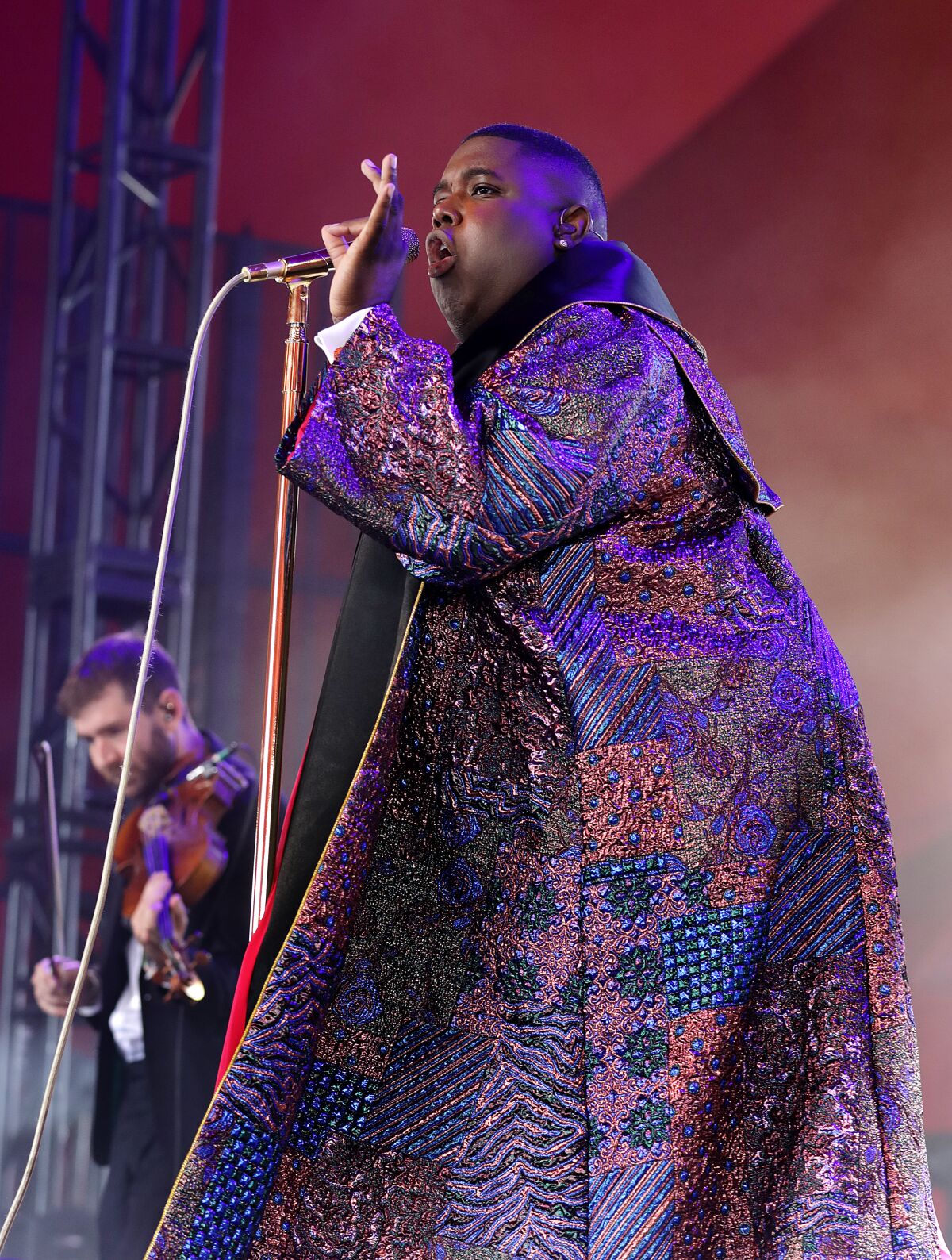 A man wearing a brocade overcoat sings into a microphone