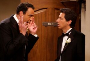 Two men in tuxedos face each other next to a men's room door